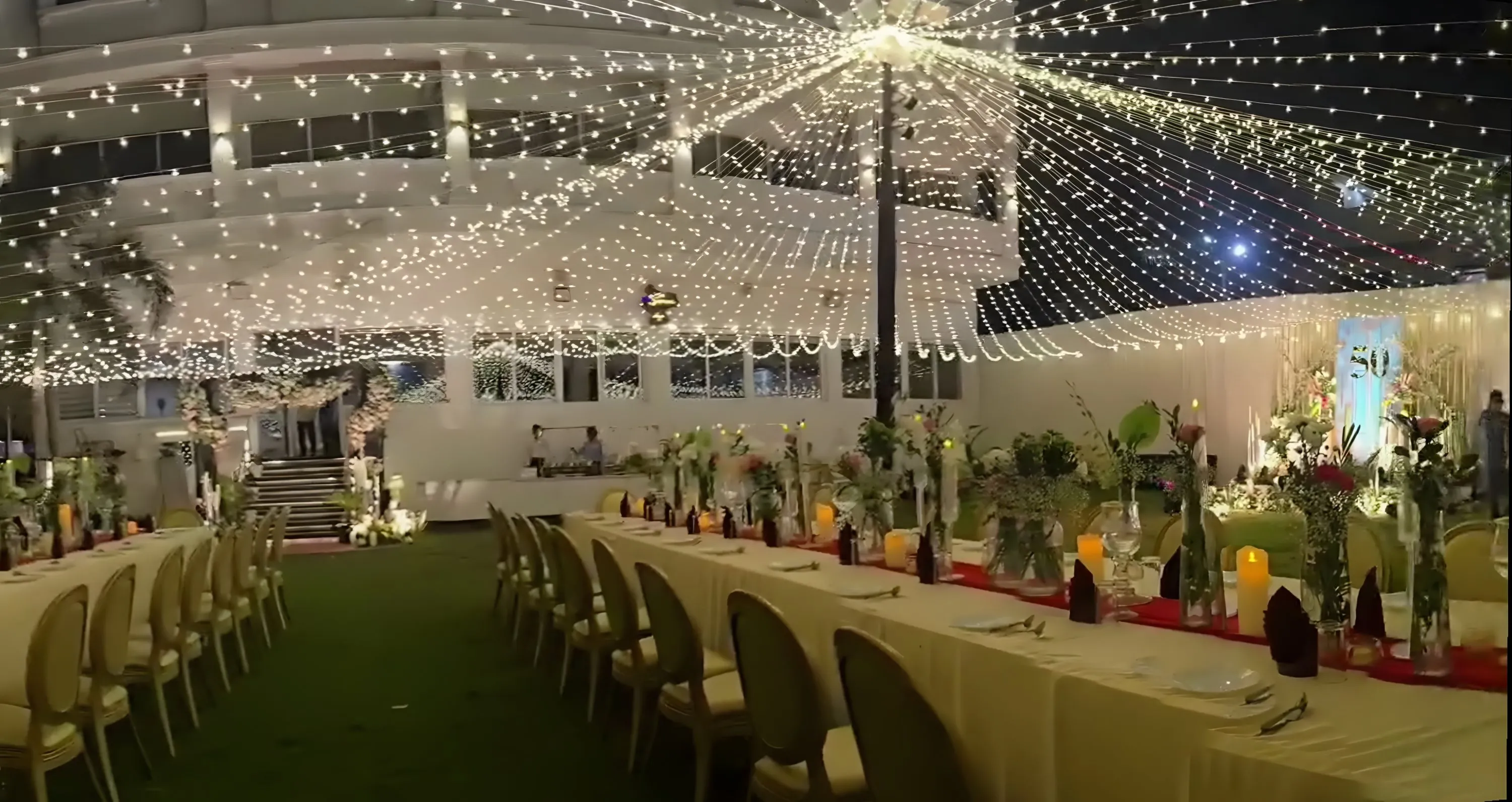 Panorama of the lawn at night: the hotel building, the floral entrance steps, the light canopy and the dinner tables